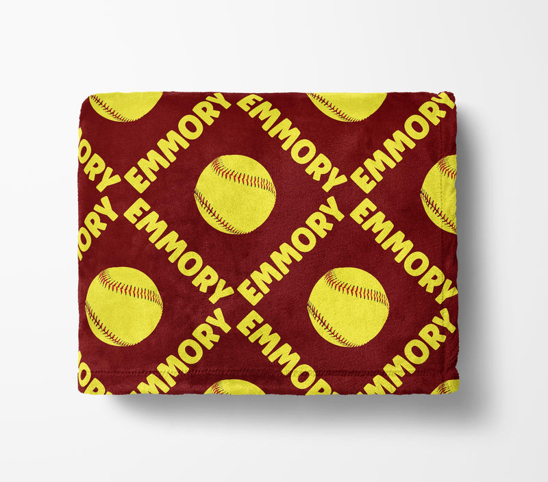 a red blanket with a repeating pattern of yellow softball images and the name "Emory" written in white.
