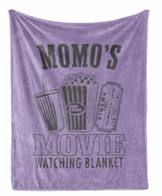 The image is a purple blanket with a graphic design that says "Momo's Movie Watching Blanket" and includes illustrations of popcorn, a soda cup, and a movie ticket.