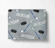 A gray blanket with a repeating pattern of hockey sticks and a puck, along with the name "JACKSON" printed on it.