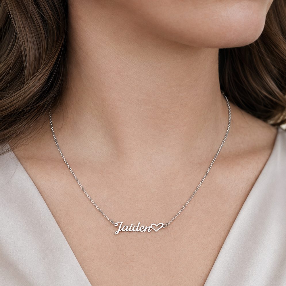 A close-up of a woman wearing a delicate silver chain necklace with a personalized nameplate that reads 'Jaiden' in cursive script, accompanied by a small heart at the end.